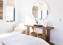 An extra-large round white mirror hangs from a white wall over a rope console table positioned in a lovely bedroom over wicker baskets.