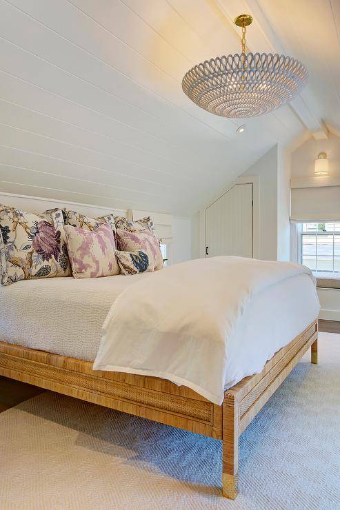 Mounted to a white shiplap vaulted ceiling, an Oly Studio Pipa Bowl Chandelier lights an attic bedroom furnished with a wood and rattan bed dressed in white bedding topped with lilac pillows.