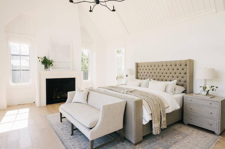 Elegant bedroom boasts a taupe tufted wingback bed placed on a gray vintage rug and positioned between gray nightstands lit by crystal lamps. An ivory wingback settee is placed at the end of the bed under a white plank vaulted ceiling. A white abstract art piece is placed atop a white fireplace mantel located between windows.