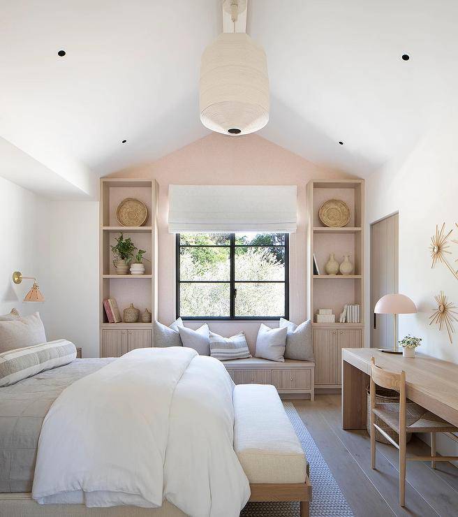Bedroom features pink wallpaper on the back of built in bookshelves over light brown reeded cabinets with a reeded bench under a window, a bed with white bedding and a wooden bench with white cushion at the foot of the bed and a long wooden desk lit by a pinkn lamp with wooden chair.