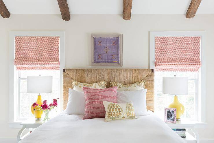 A fresh, rustic bedroom gets a lift with a woven headboard flanked by yellow lamps atop white lacquered nightstands under white and red roman shades. White and gold pillows accent white bedding delivering a boho touch to the bedroom design. A purple southwest style embroidery decorates the wall over the bed under a vaulted ceiling with rustic wood beams for dimension and style.