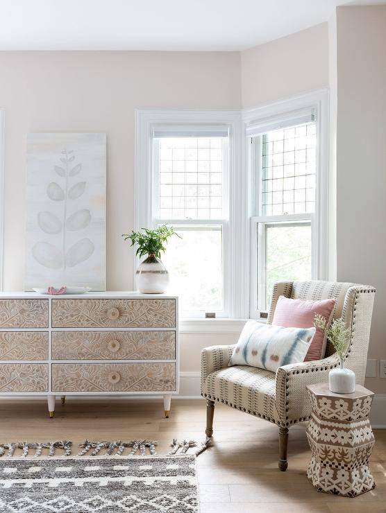 Charming bedroom features a white dresser finished with carved wood drawers and topped with a canvas art piece. A vintage burlap wingback chair is paired with a stunning carved wood accent table.