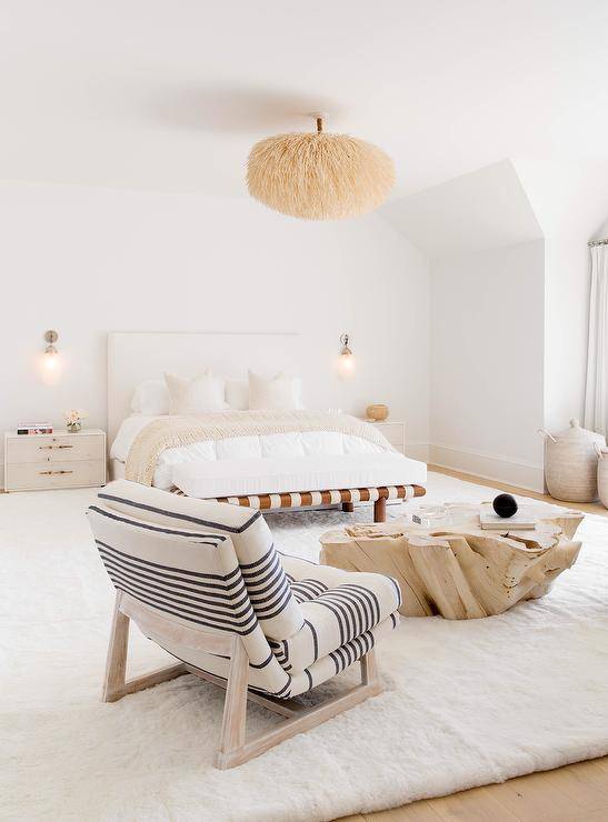 Restful neutral toned bedroom features an ivory and black striped lounge chair placed on a plush white rug at a driftwood coffee table.