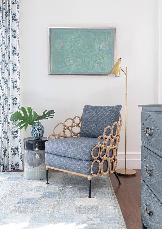 A green art piece hangs over a rattan chair accented with blue cushions and placed on a blue rug beside a blue ombre stool accent table and lit by an Aerin Clemente Floor Lamp.