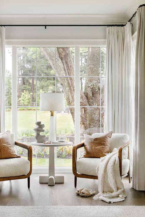 Lovely master bedroom sitting area features a round white accent table placed in front of large windows covered in white curtains and between wood and fabric vintage chairs topped with brown leather pillows.