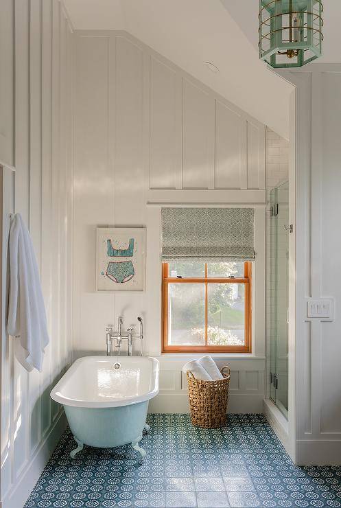 Charming white and blue cottage bathroom features a sky blue clawfoot bathtub placed on blue mosaic floor tiles in front of a polished nickel floor mount tub filler and beneath a framed blue swimsuit art piece hung from a board and batten wall. The room is illuminated by a mint green vintage lantern.