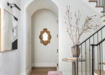 Foyer features a picture light over gray abstract art and a purple velvet skirted stool at a round gray table.