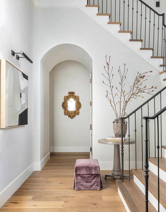 Foyer features a picture light over gray abstract art and a purple velvet skirted stool at a round gray table.