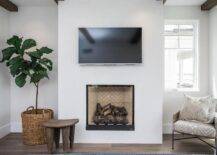 Perfect for bedrooms, this sleek fireplace has a clean black trim and transitional styling, which showcases a tan herringbone firebox under a flat panel tv mounted overhead. A wood stool and a fiddle leaf fig plant furnish the space along with a beige pattern chair accented with a chevron fringe pillow.
