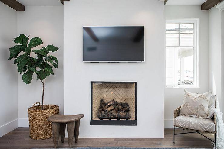Perfect for bedrooms, this sleek fireplace has a clean black trim and transitional styling, which showcases a tan herringbone firebox under a flat panel tv mounted overhead. A wood stool and a fiddle leaf fig plant furnish the space along with a beige pattern chair accented with a chevron fringe pillow.