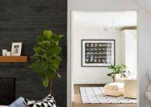 A brown chunky mantel on a black brick fireplace adds a chic contrast in a bedroom accented with a fiddle leaf fig plant. Contemporary furniture reflects a fresh style and echoes the clean-lined look of the walls styled with black and white pattern area rugs.
