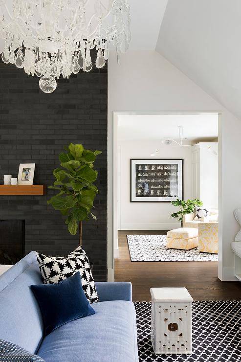 A brown chunky mantel on a black brick fireplace adds a chic contrast in a bedroom accented with a fiddle leaf fig plant. Contemporary furniture reflects a fresh style and echoes the clean-lined look of the walls styled with black and white pattern area rugs.