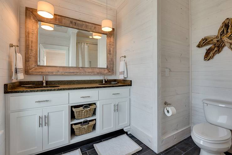 Coastal bathroom features whitewashed oak walls framing driftwood star over toilet as well as beveled wood mirror over double washstand with black countertop and small drum pendants over his and her sinks over slate tiled floor.
