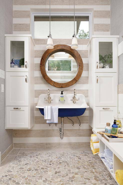 A window framed by white and gray striped wall tiles is located over a round wooden mirror hung between glass front cabinets stacked over white linen cabinets. A blue Kohler Brockway Sink floats over river rock floor tiles. A white built-in bench boasts cubbies stuffed with towels.