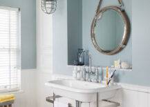 Nautical style bathroom features over the sink niche filled with a large rope bound captain's mirror illuminated by a pair of large fisherman's pendant lights hung above a chrome washstand sink with white porcelain sink and gooseneck bridge faucet. The blue cottage bathroom boasts blue upper walls, painted Zoffany Paint Dufour, over lower wall clad in tongue and groove wainscoting alongside gray tile like wood flooring which is topped with a woven nautical style basket which corrals books beside the sink.