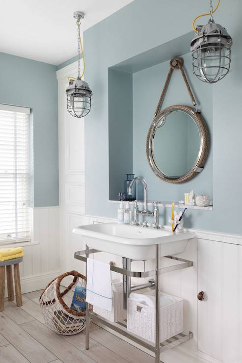 Nautical style bathroom features over the sink niche filled with a large rope bound captain's mirror illuminated by a pair of large fisherman's pendant lights hung above a chrome washstand sink with white porcelain sink and gooseneck bridge faucet. The blue cottage bathroom boasts blue upper walls, painted Zoffany Paint Dufour, over lower wall clad in tongue and groove wainscoting alongside gray tile like wood flooring which is topped with a woven nautical style basket which corrals books beside the sink.