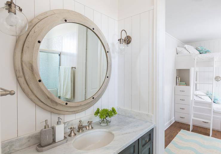 Kids' cottage bathroom features vertical shiplap walls lined with a large round nautical mirror illuminated by clear glass barn sconce over a gray distressed washstand topped with white marble fitted with an oval sink and satin nickel faucet.