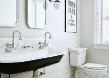 A black and white trough sink is fixed in a black and white kids' bathroom over a teak step stool placed on black and white geometric floor tiles in front of white shiplap trim. Curved nickel mirrors are lit by an oil rubbed bronze and glass globe sconce. Typography art hangs above a white porcelain toilet.