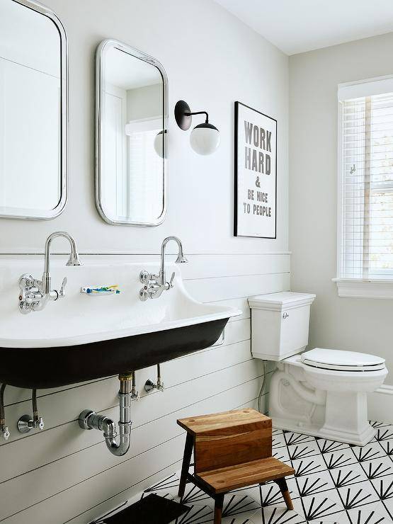 A black and white trough sink is fixed in a black and white kids' bathroom over a teak step stool placed on black and white geometric floor tiles in front of white shiplap trim. Curved nickel mirrors are lit by an oil rubbed bronze and glass globe sconce. Typography art hangs above a white porcelain toilet.