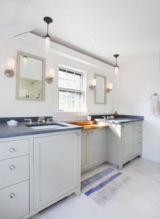Coastal bathroom with gray built-in vanity accented with brushed nickel hardware alongside gray countertops which frame undermount porcelain sinks below light gray vanity mirrors illuminated by nickel uplight wall sconces. The vanity features a drop-down console topped with butcher block below a small sash window atop gray tiled floors below layered with a striped blue bath mat.