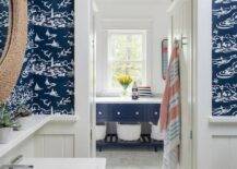 Lit by cage sconces, a round rope mirror hangs from a wall clad in navy blue nautical wallpaper lined with white shiplap. The mirror hangs over a white shelf and a white pedestal sink.