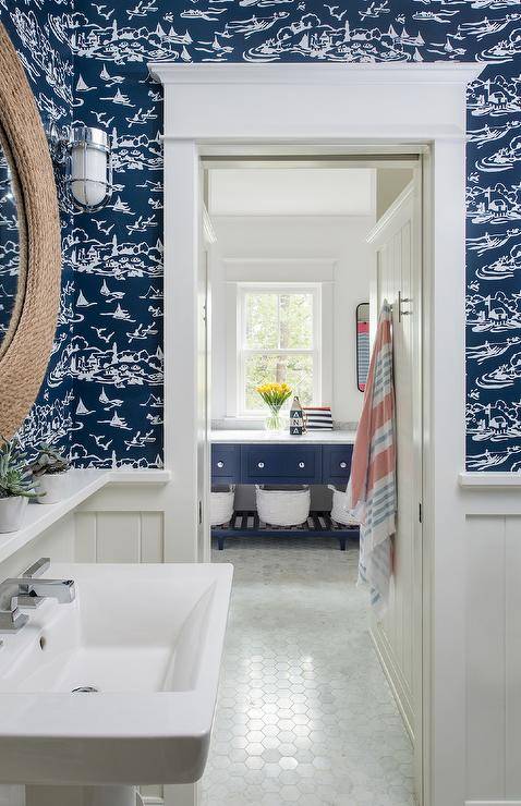 Lit by cage sconces, a round rope mirror hangs from a wall clad in navy blue nautical wallpaper lined with white shiplap. The mirror hangs over a white shelf and a white pedestal sink.