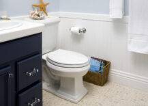 Coastal style blue bathroom with pale blue walls paired with white beadboard surround. Navy blue bathroom vanity with nickel and acrylic hardware. Beautiful pebble bathroom floor, wicker magazine rack and beachy accents.