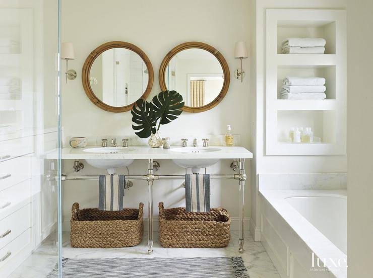 Chic coastal bathroom features a pair of round bamboo vanity mirrors illuminated by Thomas O'Brien Bryant Sconces situated over a nickel and glass dual washstand fitted with oval sinks with seagrass baskets tucked underneath alongside a gray rug layered atop marble floors. A marble top double washstand is flanked by built-ins to the left and built-in shelving over a wainscoted tub to the right.
