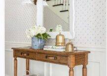 Brass sconces are mounted to a white and blue wallpapered wall lined with white wainscoting and flank a white scalloped mirror hung over a farmhouse console table.