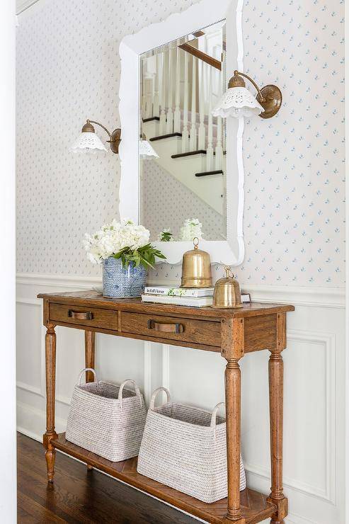 Brass sconces are mounted to a white and blue wallpapered wall lined with white wainscoting and flank a white scalloped mirror hung over a farmhouse console table.