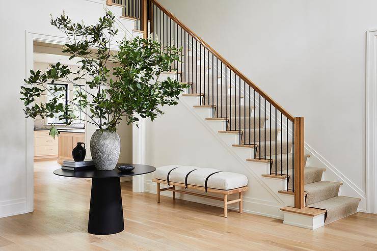 A styled round black table sits in the center of a foyer featuring a wood bench with leather straps placed against a staircase wall.