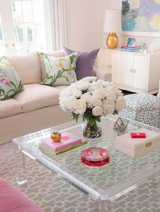 A styled square lucite coffee table sits on a gray geometric rug with pink couch and colorful decor accents