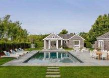 A gray shingled pool house faces an in-ground swimming pool surrounded by slate pavers and accented with white wicker loungers.