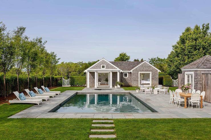 A gray shingled pool house faces an in-ground swimming pool surrounded by slate pavers and accented with white wicker loungers.