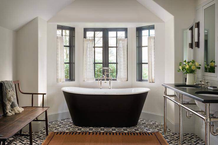 Cottage style bathroom features a bay window with black clawfoot tub on black and white mosaic floor tiles, a black top sink vanity and a wooden bench.