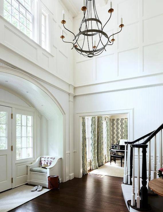 Beautifully styled two story foyer is lit by a French wrought iron chandelier hung over a dark stained wood floor contrasted with white breadboard walls fixed beneath board and batten upper walls.