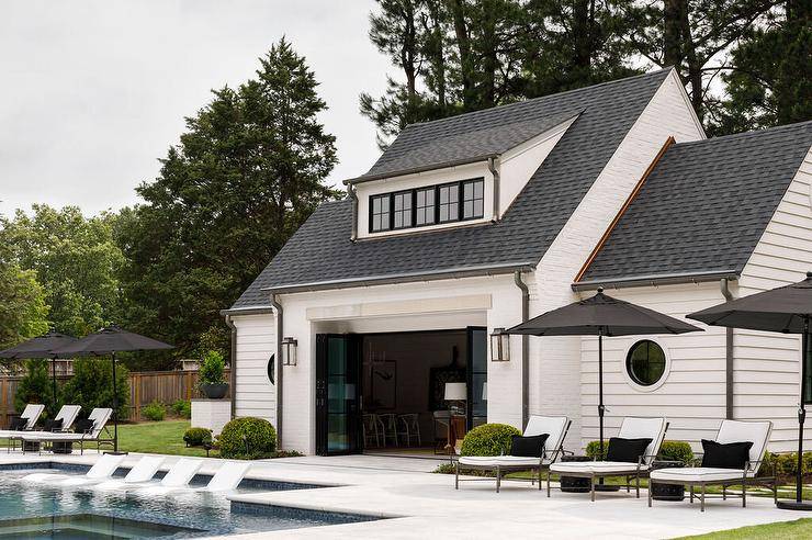 White pool house with gray roof features folding glass doors and white pool loungers under black umbrellas.