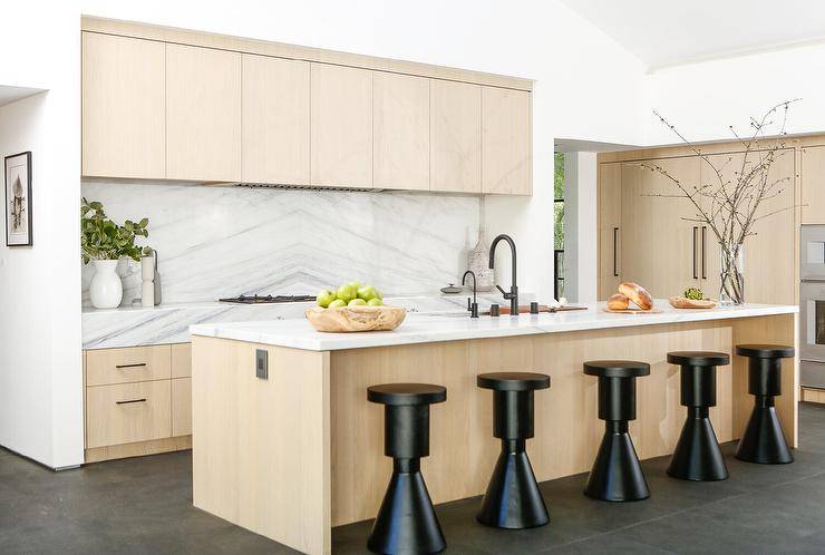 Kitchen features modern black stools at a tan center island topped with light gray honed marble with an island sink that boasts a black gooseneck faucet and tan cabinets with a light gray honed marble bookmatched kitchen backsplash.