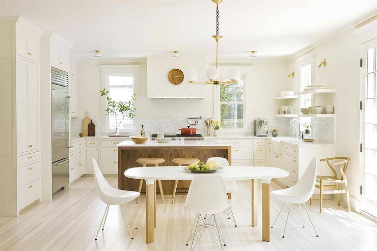 Sunny and bright, this beautiful airy white U-shaped kitchen features a white and wood decor theme. A wood stained island features quartz waterfall countertops around white cabinetry with brass hardware and white floating shelves. Backless wood counter stools sit at the island providing seating along with a midcentury modern kitchen table with round edges and wood dipped legs. White molded plastic chairs frame the table on blond oak wood floors under white vintage enamel kitchen flush-mount lights and a white glass and brass chandelier. Stainless steel appliances are an upgrade in this open concept kitchen including a refrigerator, cooktop stove and swivel swing arm pot filler.