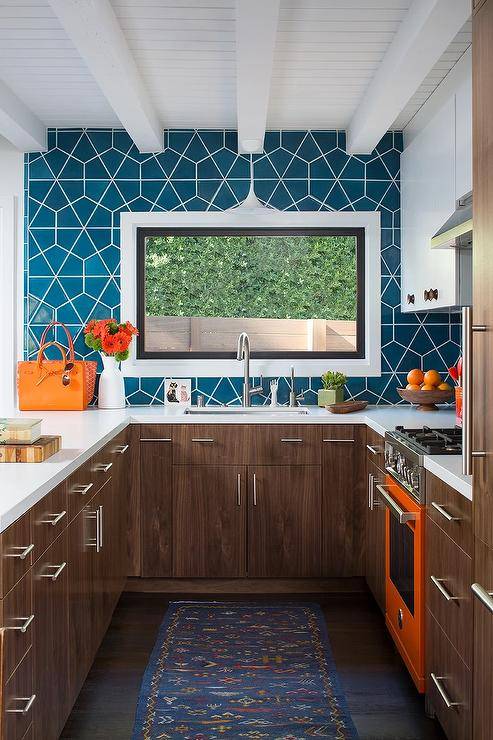 Charming brown, orange, and blue u-shaped kitchen boasts an orange stove placed between brown veneer cabinets donning satin nickel hardware. A picture window is framed by blue geometric wall tiles and located beneath a white plank ceiling and over a sink iwth a satin nickel gooseneck faucet.