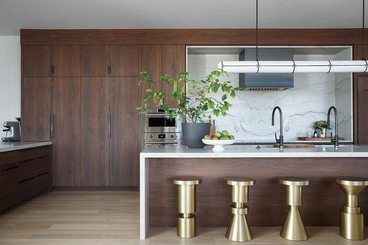 A brown veneer island topped with a marble waterfall countertop is matched with modern brass backless stools and completed with a sink and oil rubbed bronze gooseneck faucet illuminated by a black and white linear chandelier.