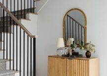 A tan wood bar cabinet accented with a crystal lamp is placed to the side of a staircase and beneath an arch bamboo mirror.