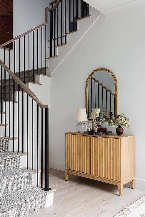 A tan wood bar cabinet accented with a crystal lamp is placed to the side of a staircase and beneath an arch bamboo mirror.