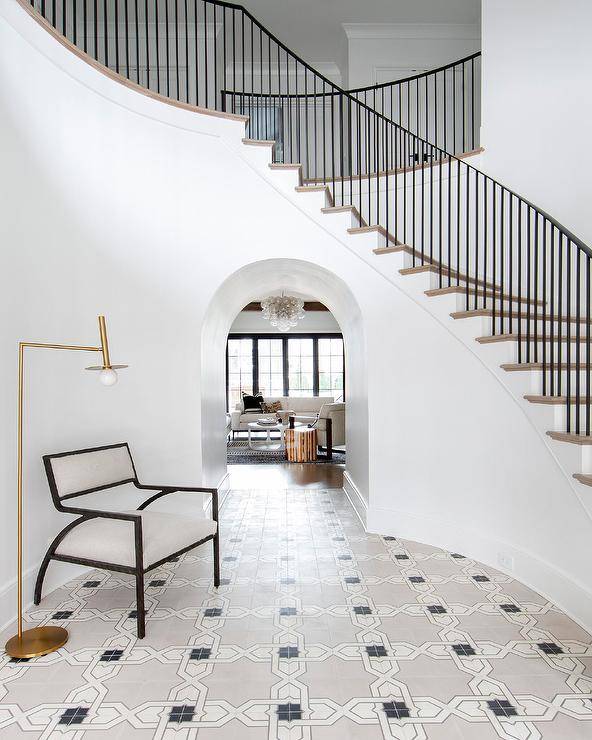 A rotunda foyer features black and white trellis floor tiles and an off white and wooden chair lit by a brass floor lamp.