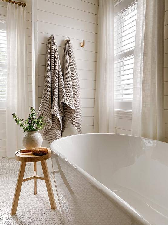 Modern brass towel hooks are fixed to a shiplap wall in a cottage bathroom featuring windows covered in white shutters and sheer white curtains.
