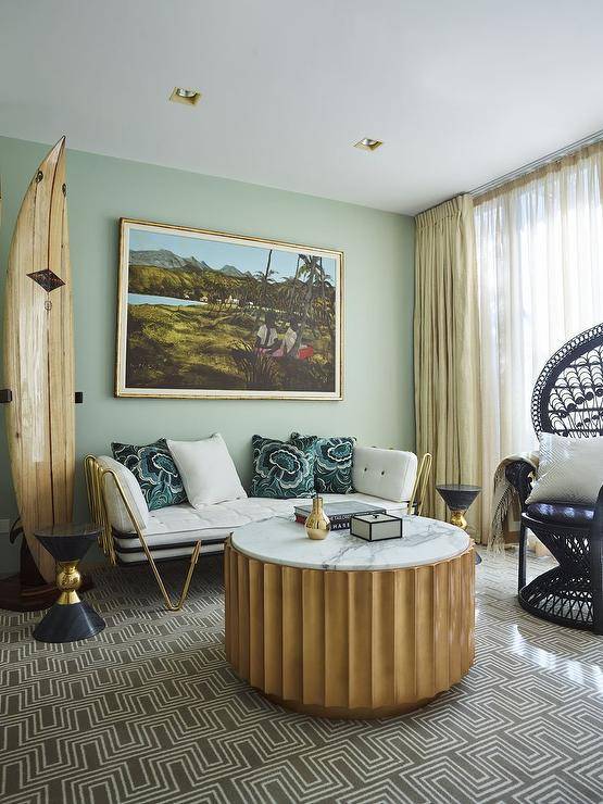 Living room features a round fluted coffee table with marble top on a tan rug, a brass and white daybed accented with blue pillows and a decorative surfboard.
