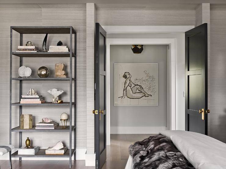 Black double bedroom doors with brass door knobs invite into a bedroom furnished with a twin bed and a black metal bookcase styled with decor, magazines, and books. Platinum gray grasscloth wallpaper brings texture with a timeless natural appeal.