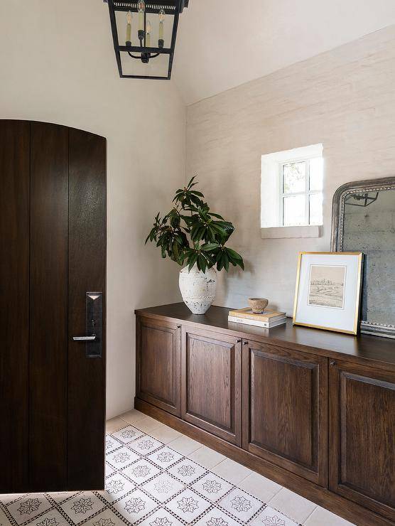 An arched brown wooden door opens to mosaic floor tiles accenting a foyer boasting a built-in credenza placed under a window and topped with a silver leaf beaded mirror.