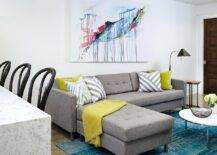 Contemporary basement family room boasts a large colorful abstract canvas art piece hung on white walls above a gray tufted sectional topped with citron yellow and gray diagonal striped accent pillows as well as a citron yellow throw blanket. In front of the sofa, a turquoise overdyed rug is positioned beneath a chrome and marble round coffee table as beside the sofa the look is completed with a clover accent table lit by a black and gold floor lamp.