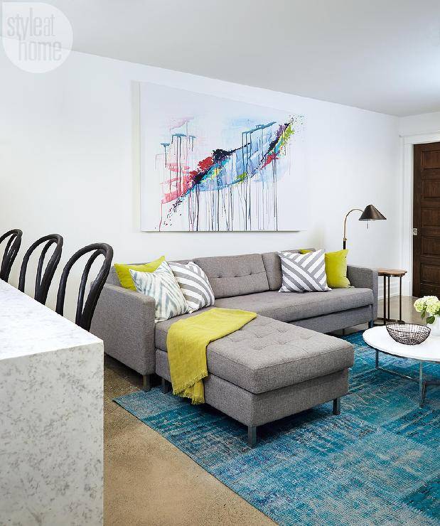 Contemporary basement family room boasts a large colorful abstract canvas art piece hung on white walls above a gray tufted sectional topped with citron yellow and gray diagonal striped accent pillows as well as a citron yellow throw blanket. In front of the sofa, a turquoise overdyed rug is positioned beneath a chrome and marble round coffee table as beside the sofa the look is completed with a clover accent table lit by a black and gold floor lamp.